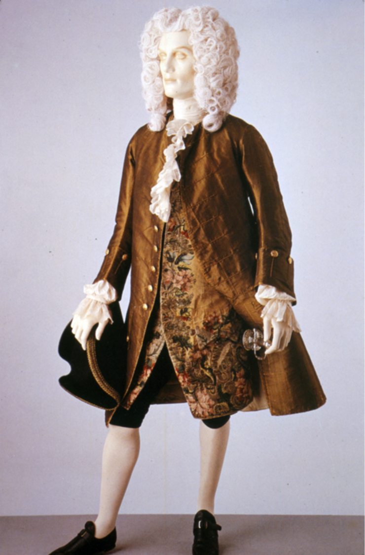 “This coat and waistcoat illustrate formal daywear for men in the 1740s. The fabric of the coat is a rich shot green and black silk. By the 1740s the waistcoat is shorter in length than the coat. It is made of yellow silk brocaded with coloured silk and silver threads. Comprised of large flowers and leaves densely covering the fabric, the brocaded pattern is typical of Late Baroque design. The coat is collarless. It fits tightly to the body, but has very full skirts pleated to the sides at the hip. The sleeve cuffs are wide, reaching about half way to the elbow. Typical of the early 18th century, the waistcoat is also sleeved, although this style was beginning to go out of fashion by the 1740s.” Victoria & Albert Museum, 1745